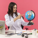 young woman doctor in white coat with stethoscope around her neck holding globe looking with smile on face sitting at the table with laptop over pink background