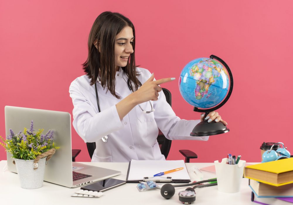 young woman doctor in white coat with stethoscope around her neck holding globe looking with smile on face sitting at the table with laptop over pink background